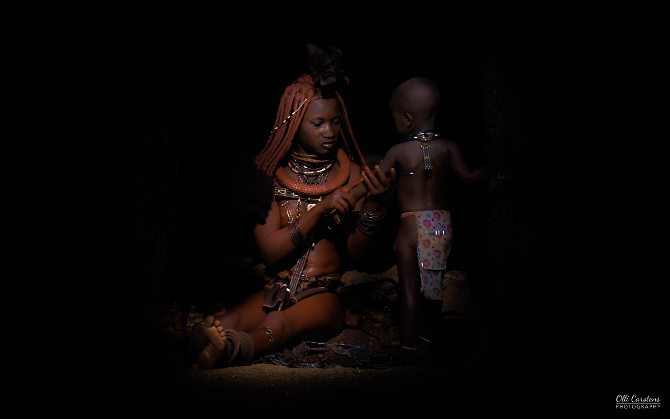 Himba-Mutter mit Kind, aufgenommen während einer Fotosafari mit Buschmann Safaris. Ein Mensch in traditioneller Kleidung sitzt neben einem Kleinkind in einem dunklen Raum.